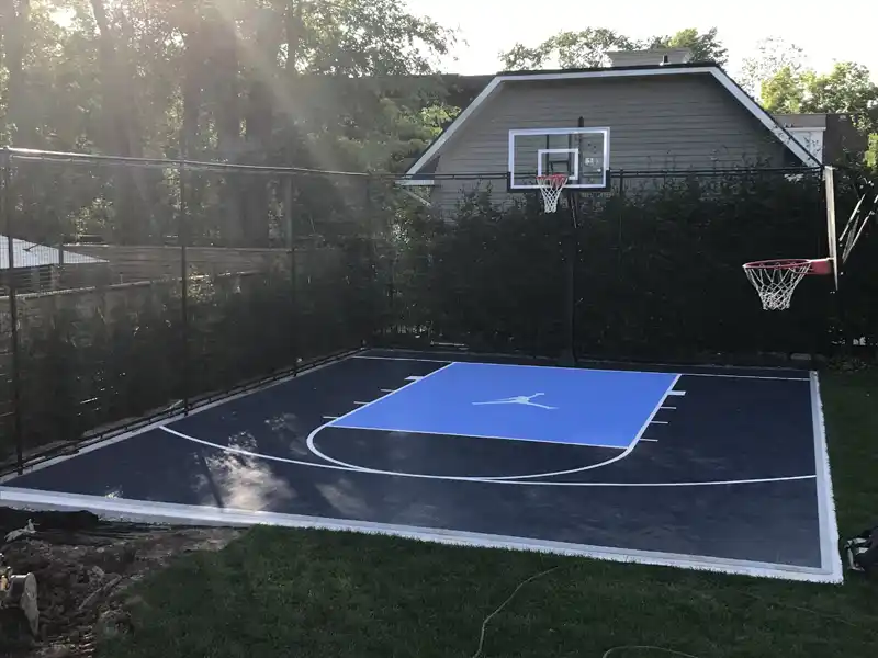 Small backyard basketball court with black SnapSports® tiles and a blue key. Features dual hoops, containment fencing, and a sleek white line layout.